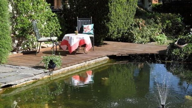 Una terraza de madera con mesa y sillas, un pequeño estanque con fuente, y abundante vegetación alrededor.