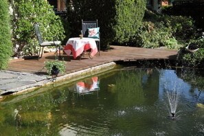 Eine Holzterrasse mit Tisch und Stühlen, ein kleiner Teich mit Springbrunnen und umgebende Bepflanzung.