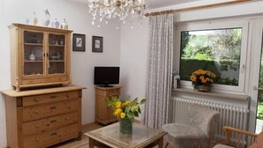 Una sala de estar con un aparador de madera, televisor, una mesa de vidrio, un jarrón floral, alfombra estampada y una ventana con cortinas.