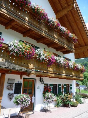 A traditional chalet with wooden balconies adorned with colorful flowers, a wooden door, and a sign indicating a restaurant. - Ferienwohnung Kneifelspitze für 2-6 Personen, 88 qm (Marktschellenberg)