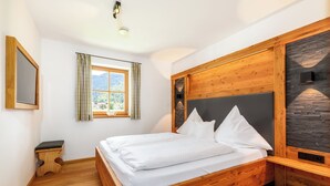 Une chambre avec un lit en bois, un petit tabouret, une fenêtre avec des rideaux et un mur en pierre servant d’élément décoratif.