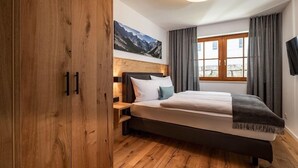 A bedroom with a wooden wardrobe, a bed with white bedding, a mountain view through the window, and a flat-screen TV on the wall.