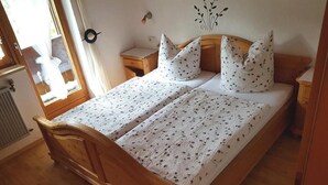 A wooden bed with a floral bedspread and matching pillows, a wooden nightstand, and a wall-mounted clock. - Apartment Baumgart for max. 3 persons, 35 sqm, 1 bedroom, balcony (Schönau am Königssee)