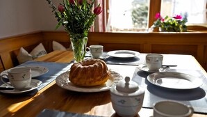 Een eettafel gedekt met borden, kopjes, een theepot en een tulbandtaart, met een bloevas en een raam op de achtergrond.