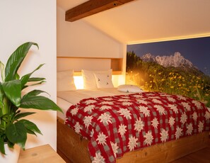 A bedroom with a bed, pillows, a red and white patterned blanket, a wooden headboard, a wall-mounted painting of a mountain, and a potted plant. - Ferienwohnung (6) (Reit im Winkl)