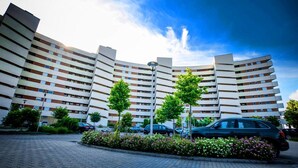 En bygning med flere etager og en buet facade, en parkeringsplads med biler samt en anlagt have med træer og blomster.