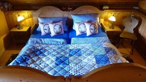 A wooden cabin room with a bed featuring a blue quilt and two pillows with clock designs. There are two bedside lamps and a wooden chair.