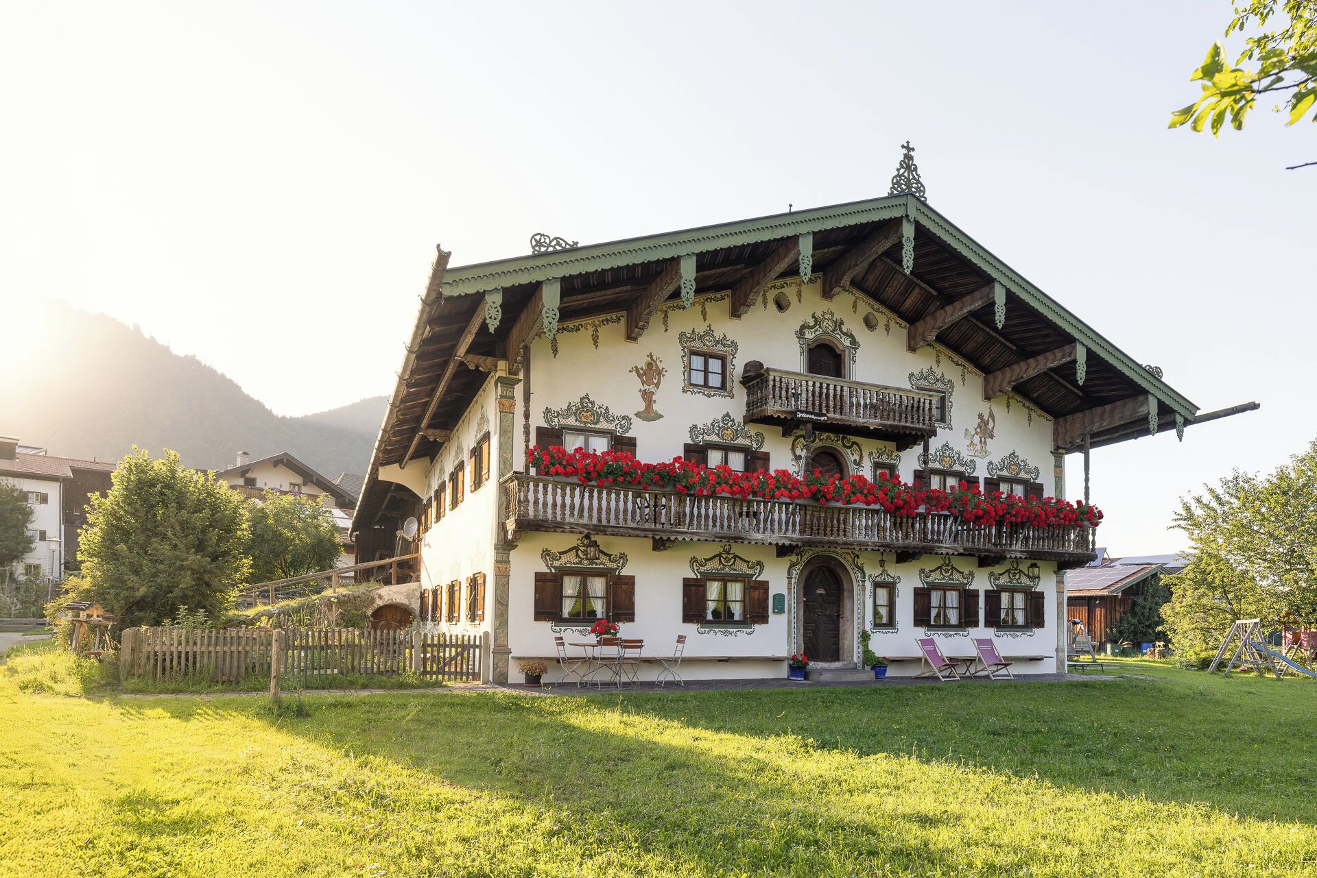 Un chalet alpin traditionnel doté d’un balcon orné de fleurs rouges, d’une clôture en bois et offrant une vue sur la verdure et les montagnes environnantes.