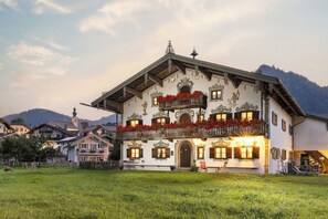 Ein traditionelles Gebäude im alpinen Stil mit einem Balkon, der mit roten Blumen geschmückt ist, einer Holztreppe und einem Blick auf die Berge.