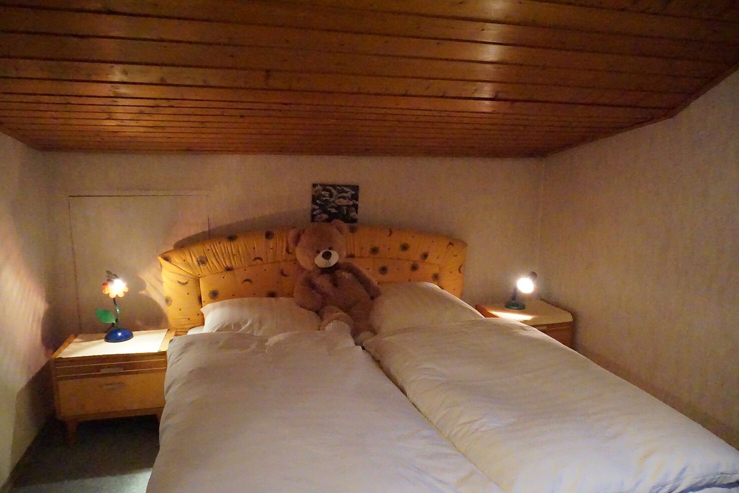 Quarto com teto de madeira, cama com roupa de cama branca, um urso de pelúcia na cama e duas luminárias de cabeceira.
