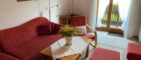 Ett vardagsrum med en röd soffa, ett träbord med en vas med gula blommor, en röd fåtölj och ett litet träbord. På väggen hänger två tavlor, det finns en golvlampa och ett fönster med gardiner.