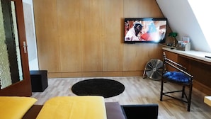 Ett mysigt rum med en tv som hänger på väggen, en trägarderob, en fläkt och en blå stol.