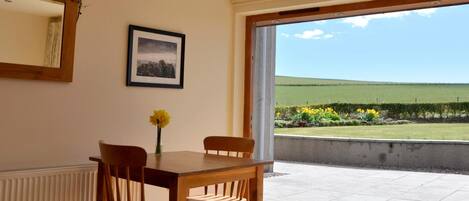 A dining area with a table and chairs, a framed picture on the wall, and a view of a green field through a large window.