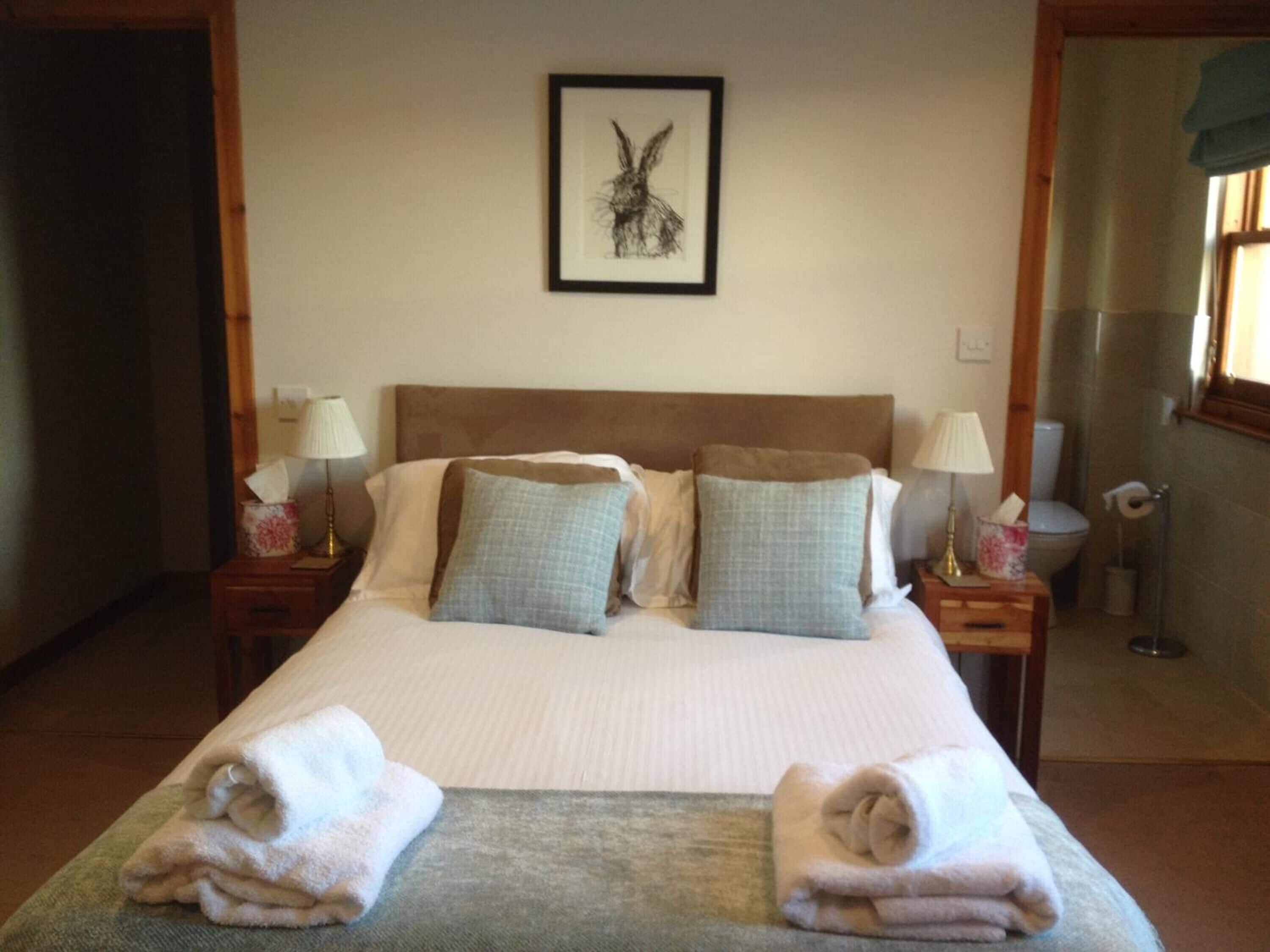 Una cama bien tendida con almohadas y toallas dobladas, un cuadro en la pared y un baño visible a través de una puerta abierta.