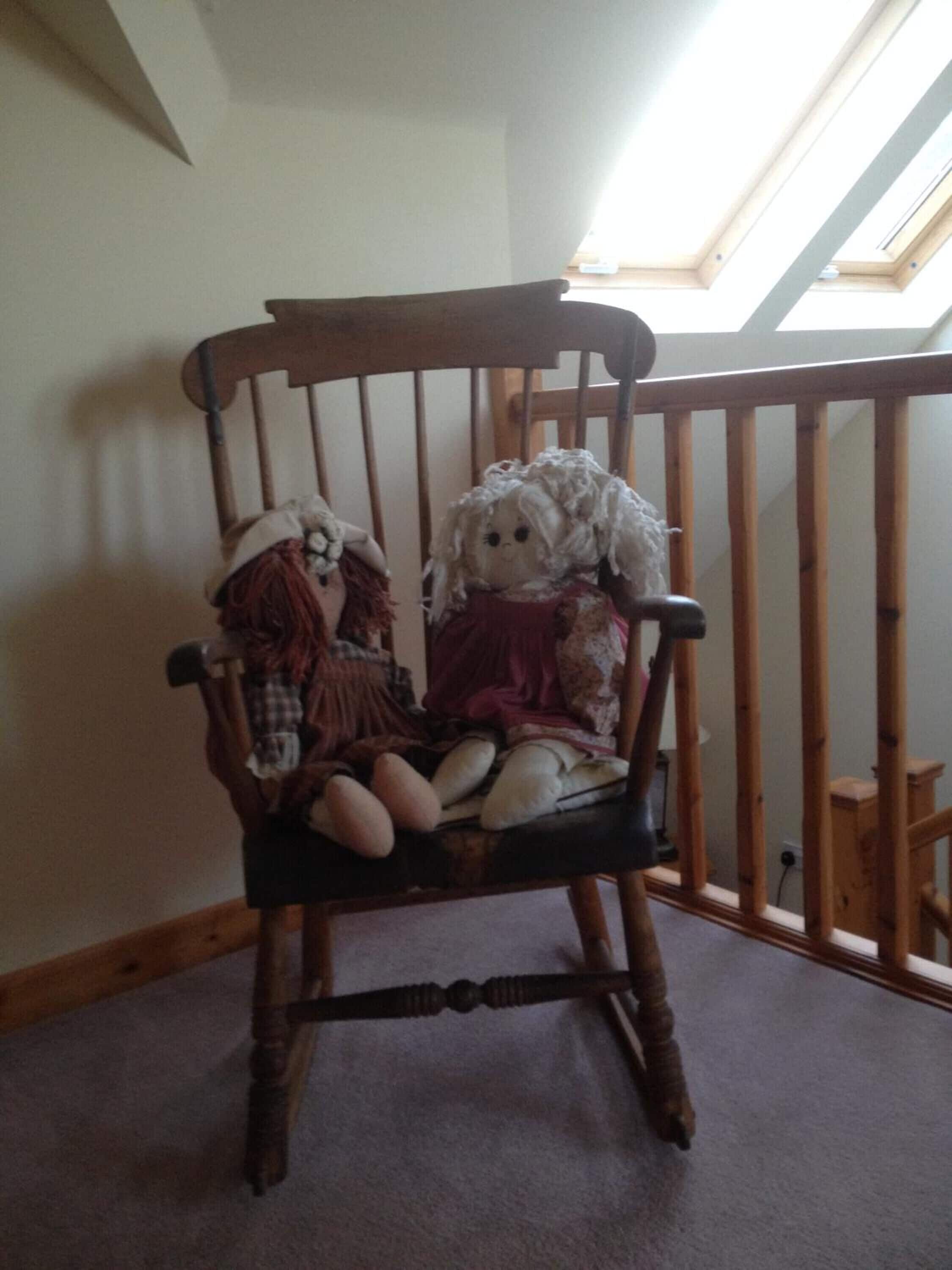A wooden chair with two dolls in a room with a sloped ceiling and a skylight.