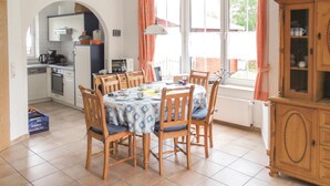 Een eetruimte met een tafel gedekt voor zes personen, omringd door houten stoelen met blauwe kussens. Op de achtergrond is een keuken met witte kasten en een magnetron zichtbaar.