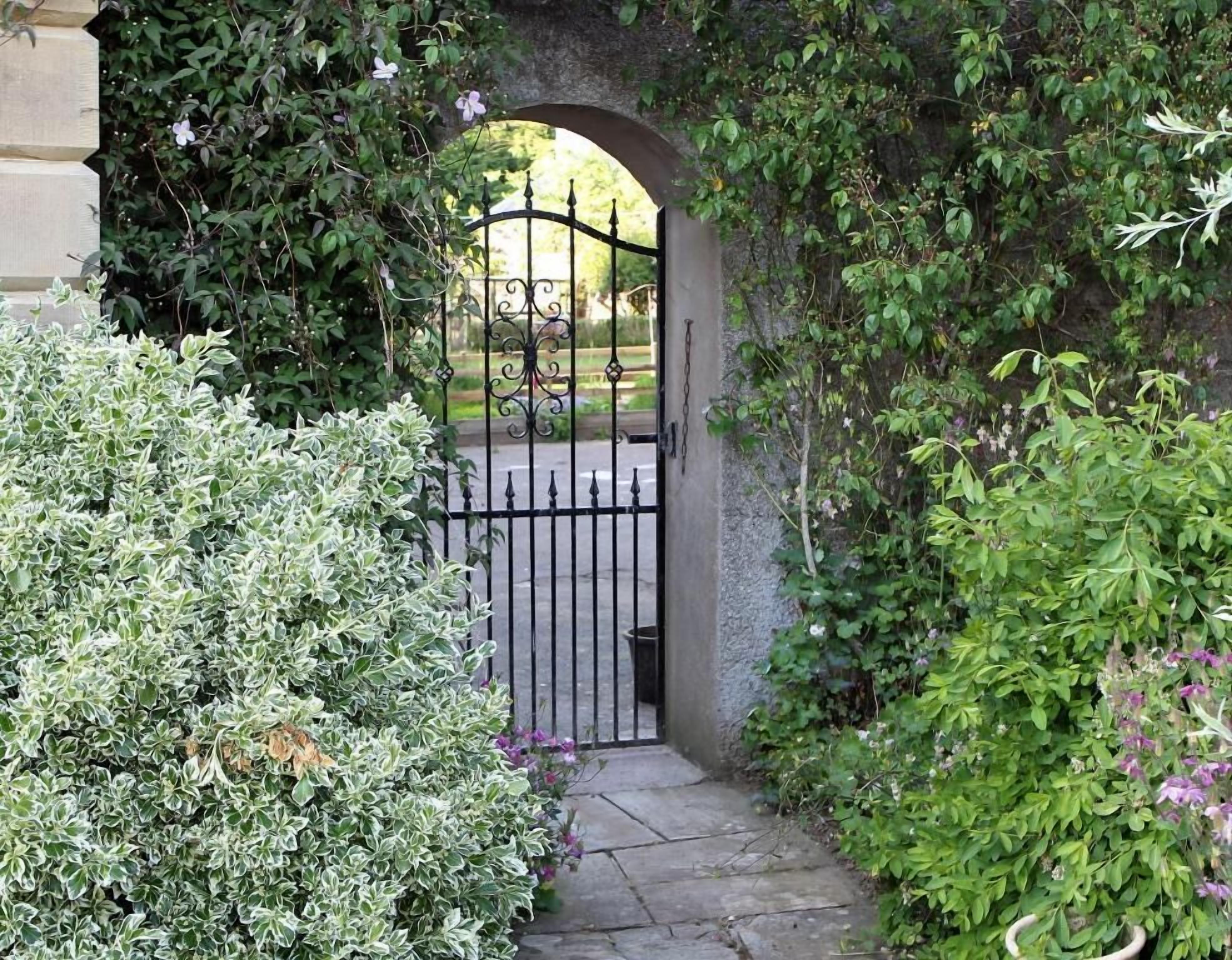 Une entrée fermée par un portail, avec un chemin de pierre menant à travers une arche, le tout entouré d’une végétation luxuriante et de plantes en fleurs.