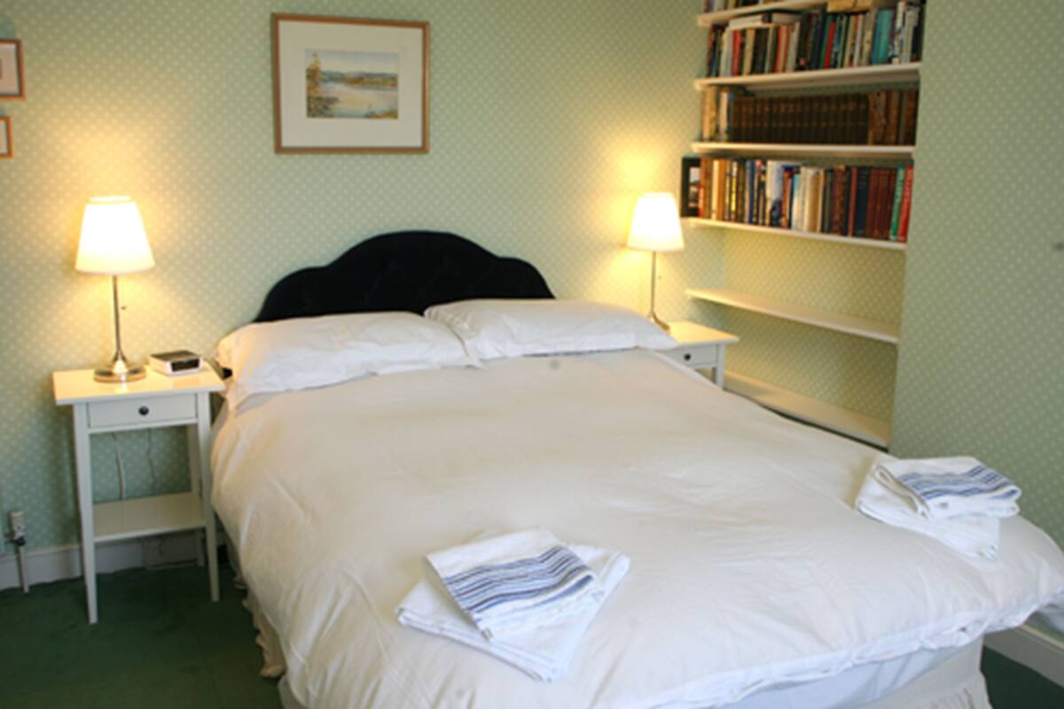 A neatly made bed with white linens and towels, two bedside lamps, a nightstand with a phone, and a bookshelf with books.