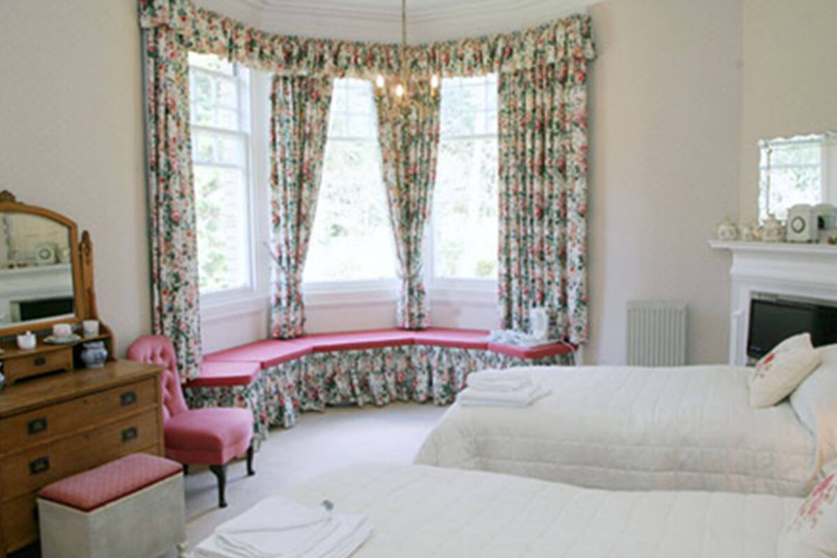 A bedroom with a bed, a dresser, a chair, a window with floral curtains, and a fireplace.