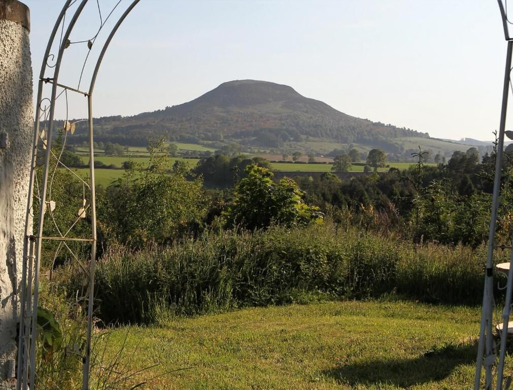 Paisaje rural con una colina al fondo, un campo cubierto de hierba en primer plano y una estructura metálica a la izquierda.