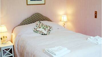 Una cama bien hecha con un cojín floral, dos lámparas de noche y toallas dobladas sobre una mesita de noche blanca.