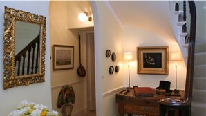 A hallway with a staircase, a mirror, framed artwork, a desk with a lamp, and a vase of flowers. - Kirklands Bed & Breakfast (Melrose)