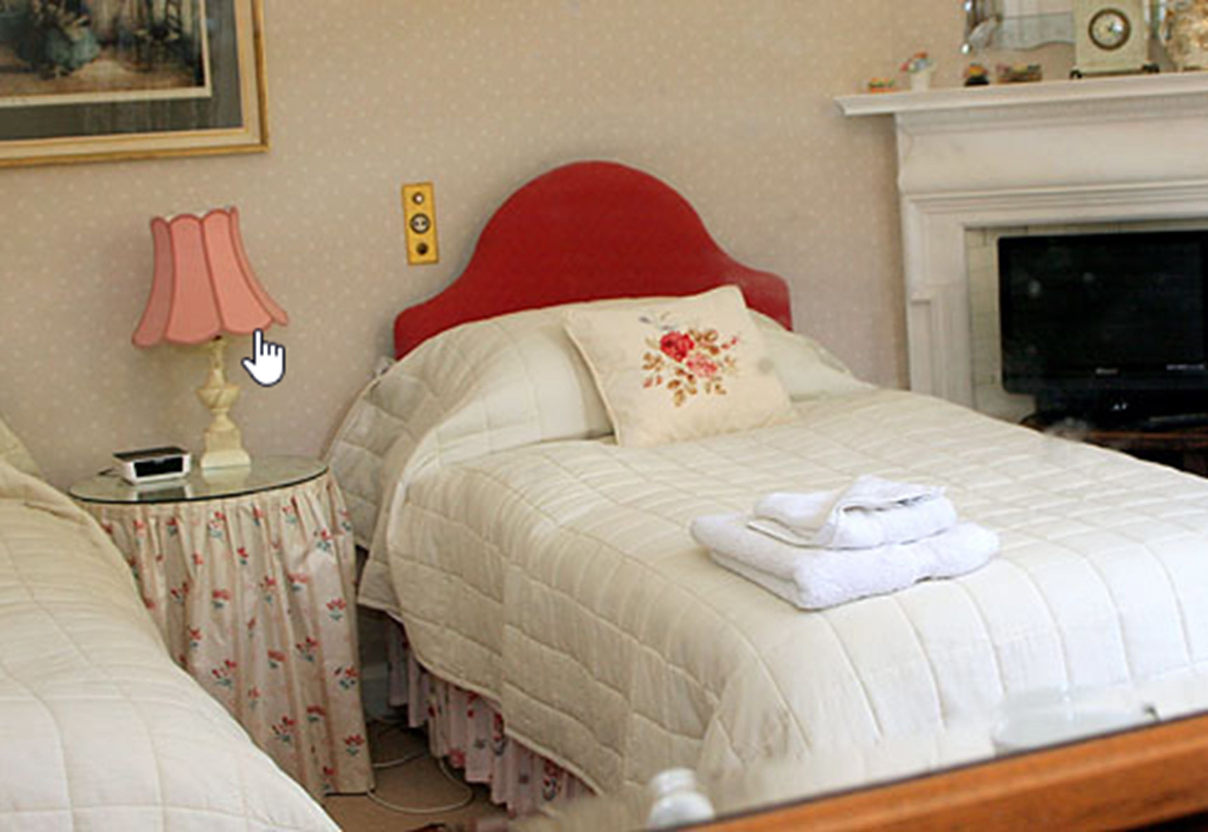 Un lit bien fait, avec une tête de lit rouge, une table de chevet surmontée d’une lampe et une nappe à motifs floraux.