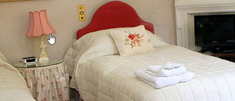 Un lit bien fait, avec une tête de lit rouge, une table de chevet surmontée d’une lampe et une nappe à motifs floraux.