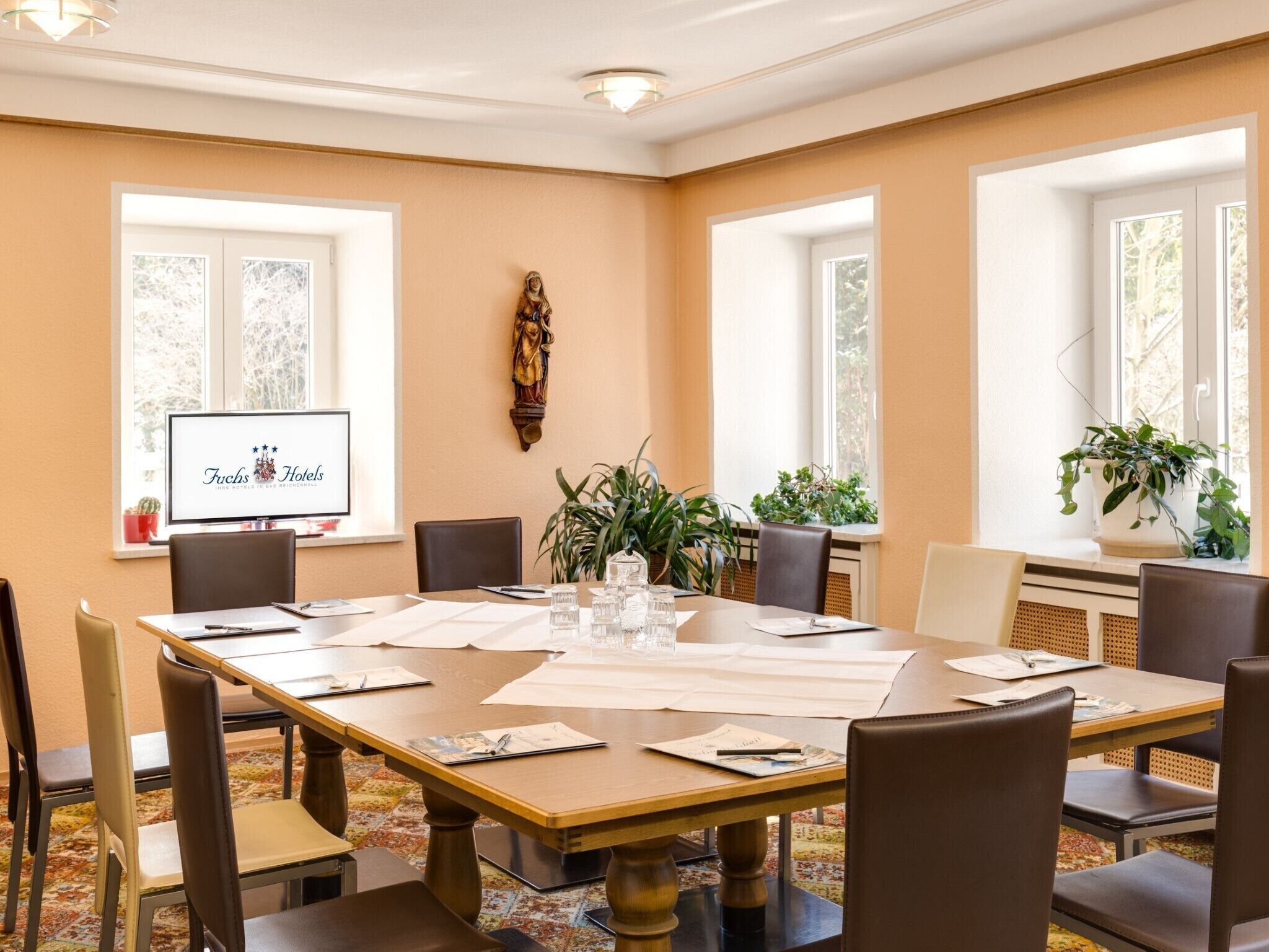 A conference room with a large table, chairs, a TV screen displaying 'Fuchs Hotels', and large windows with plants.