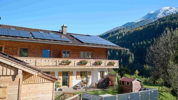 A wooden house with a balcony, solar panels, and a mountain in the background.