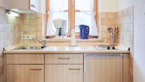 A compact kitchen with wooden cabinets, a double sink, and a window with sheer curtains. - (202) Ein-Raum-Ferienwohnung (Reit im Winkl)