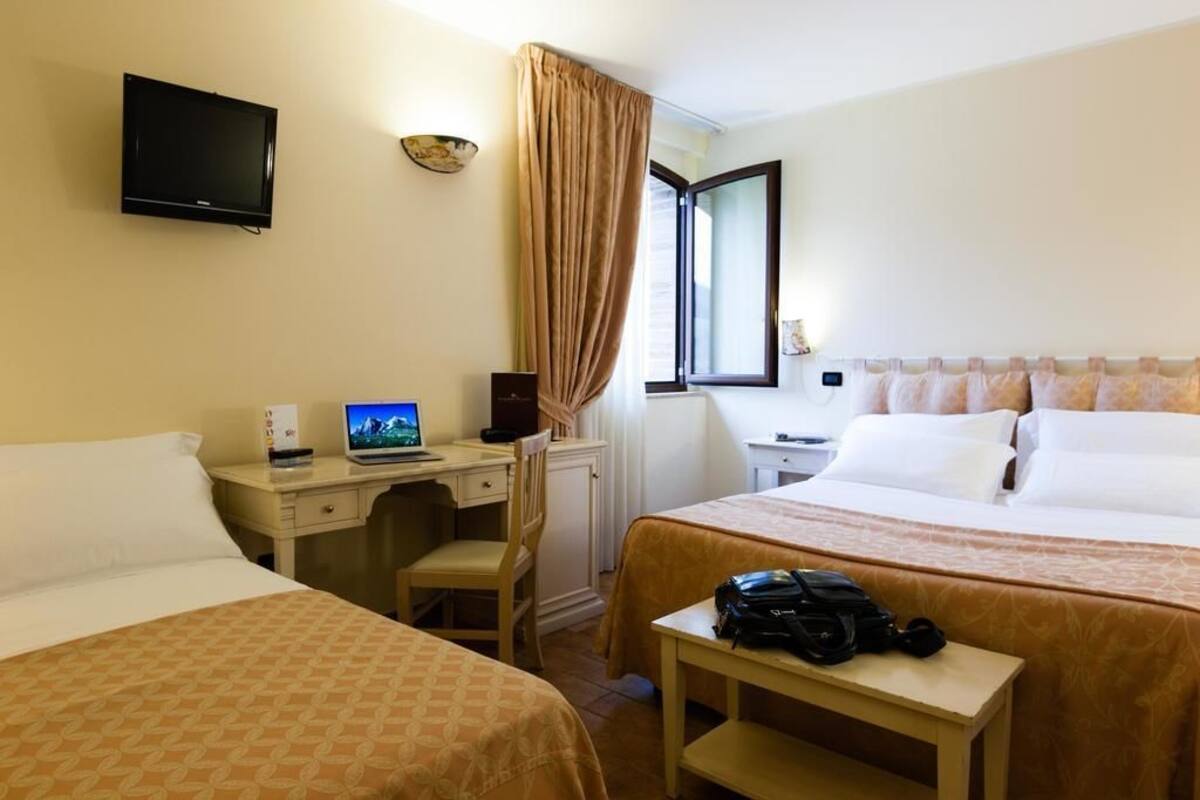 A hotel room with two beds, a desk with a laptop, a TV mounted on the wall, and a window with curtains.