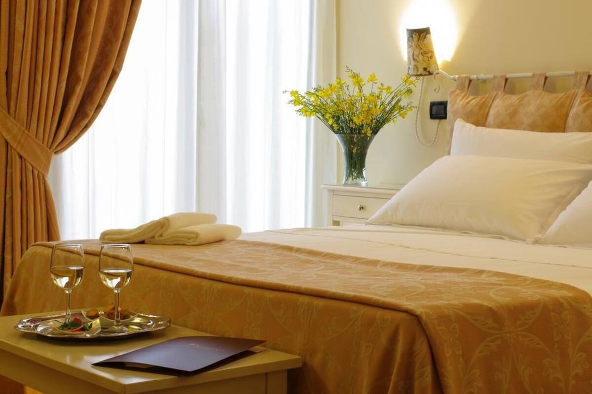 A hotel room with a bed, a bedside table with a tray of food and two glasses of water, a vase with yellow flowers, and a lamp on the wall.