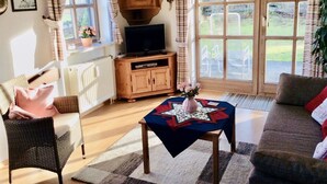 Een gezellige woonkamer met een televisie op een houten kast, een rieten fauteuil, een bank en een tafel met een bloevas.