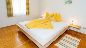 Une chambre avec un plancher en bois, un lit recouvert de draps blancs et agrémenté de plusieurs coussins jaunes, une fenêtre avec des rideaux jaunes et une petite table de chevet avec une lampe.