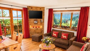 Una sala de estar con piso de madera, un sofá con cojines rojos, una mesa de centro con un jarrón de flores y un televisor montado en una pared de madera.