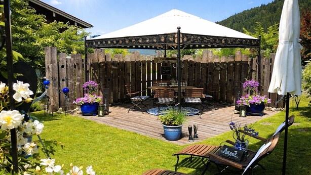 Una terraza de madera con un área de estar cubierta, rodeada por una valla de madera y macetas con plantas.