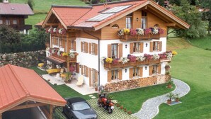 Un chalet de dos pisos con techo inclinado, balcones de madera con jardineras y un camino de entrada pavimentado, con un coche estacionado al frente.