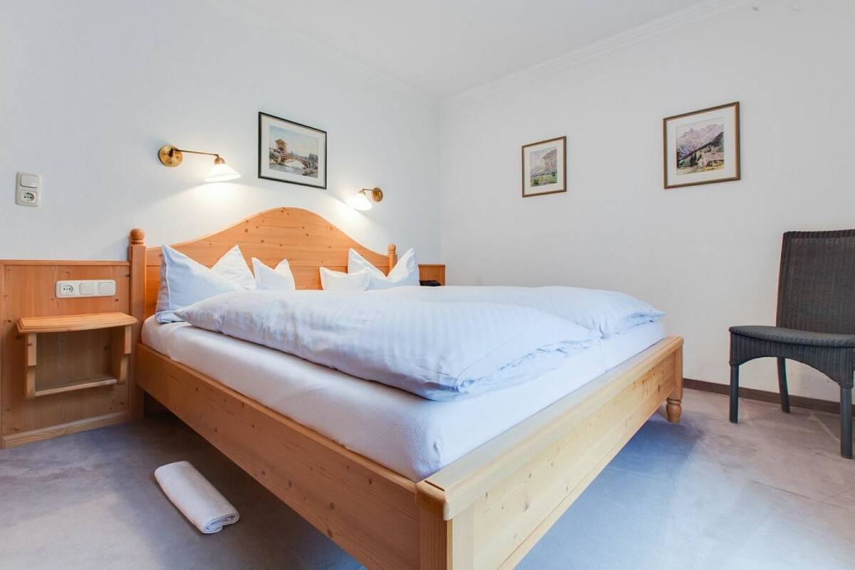 A wooden bed with white bedding, a nightstand, a chair, and two wall-mounted lamps.