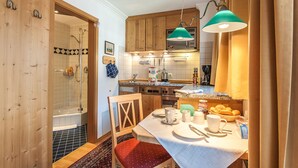 Cocina compacta con armarios de madera, una mesa de comedor con tazas y platos, y una zona de ducha visible a través de una puerta abierta.