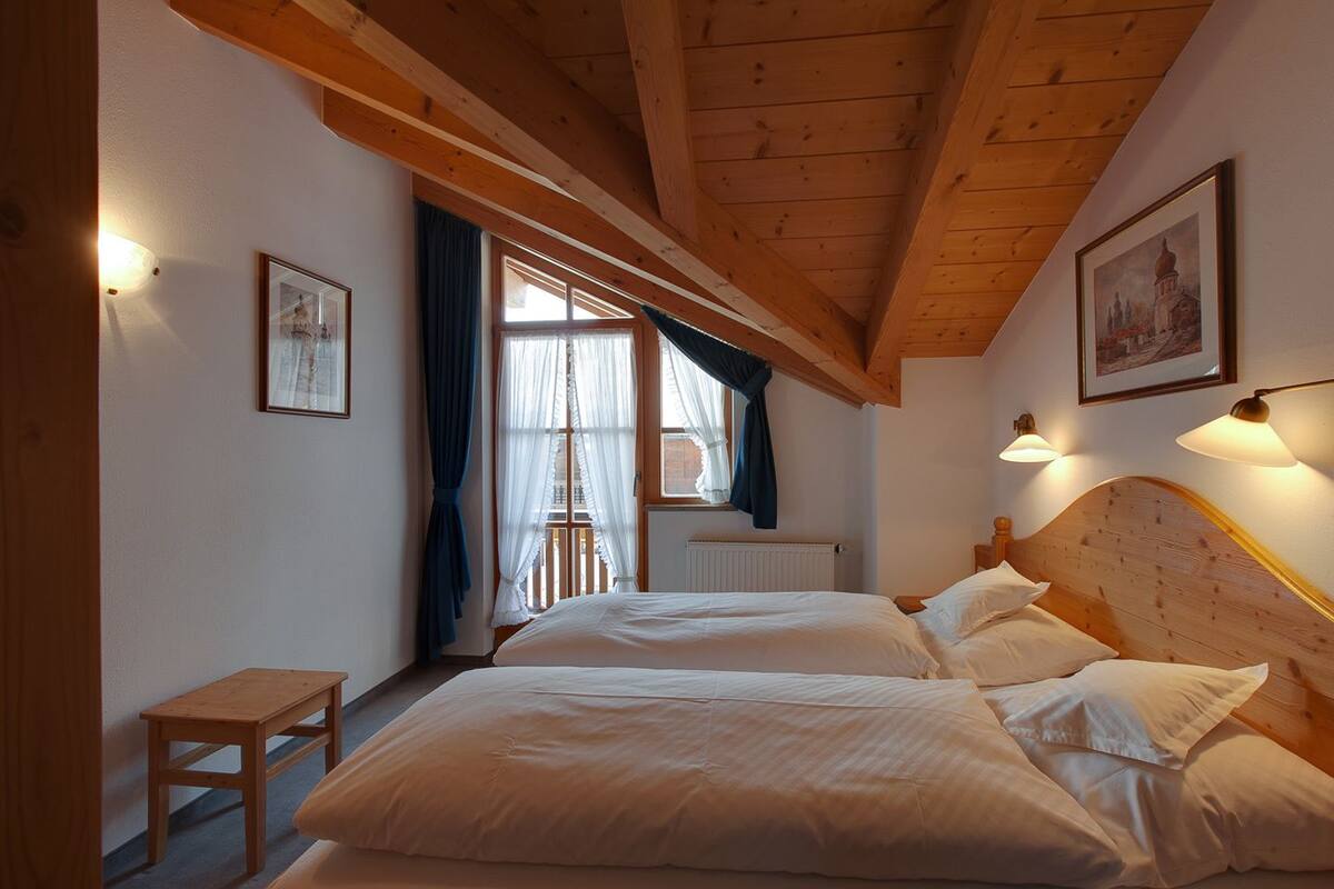 A bedroom with two beds, wooden beams, a window with curtains, and wall-mounted lights.