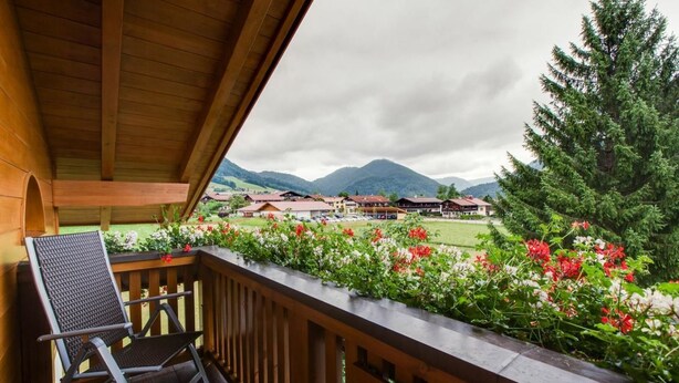 Un balcón de madera con una silla, con vistas a un paisaje montañoso con edificios y un árbol grande.