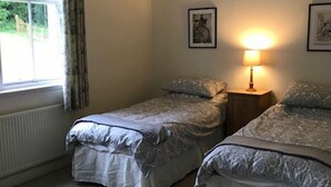 A bedroom with two single beds, a window with curtains, a bedside table with a lamp, and framed pictures on the wall. - Kirklands Cottages (Melrose)