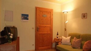 A cozy living room with a wooden door, a green sofa with cushions, a small table with a bottle, and a floor lamp. - Ferienwohnung Jenner für 1-3 Pers., ca. 34 qm, Schlafzimmer, Wohnküche, Balkon (Bischofswiesen)
