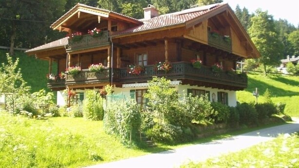 A traditional wooden chalet with a balcony, surrounded by lush greenery and a mountainous backdrop. - Ferienwohnung Jenner für 1-3 Pers., ca. 34 qm, Schlafzimmer, Wohnküche, Balkon (Bischofswiesen)