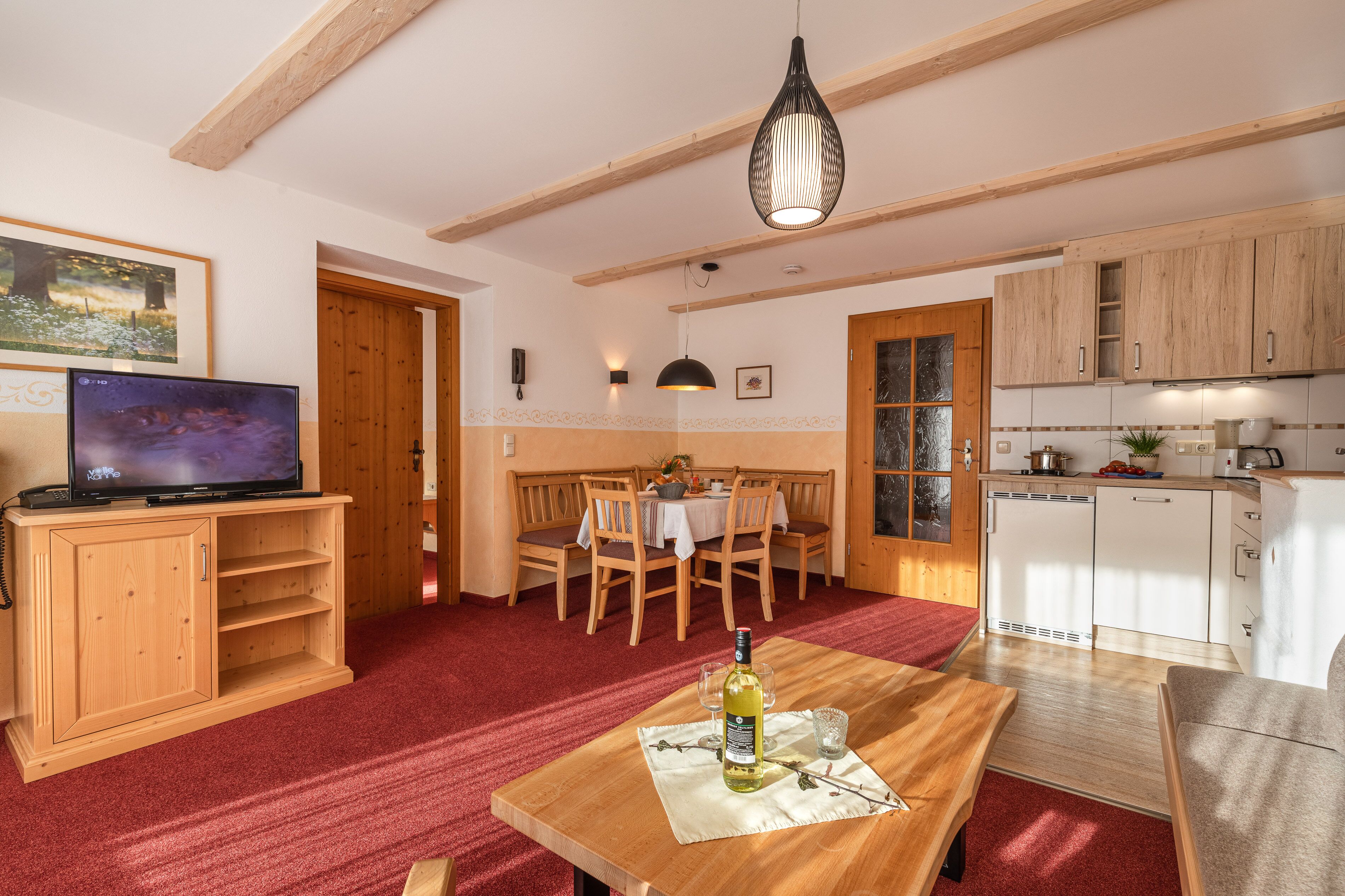 Uma sala de estar acolhedora com televisão, móveis de madeira, um espaço de jantar com mesa e cadeiras, e uma cozinha com armários brancos e luminária pendente.