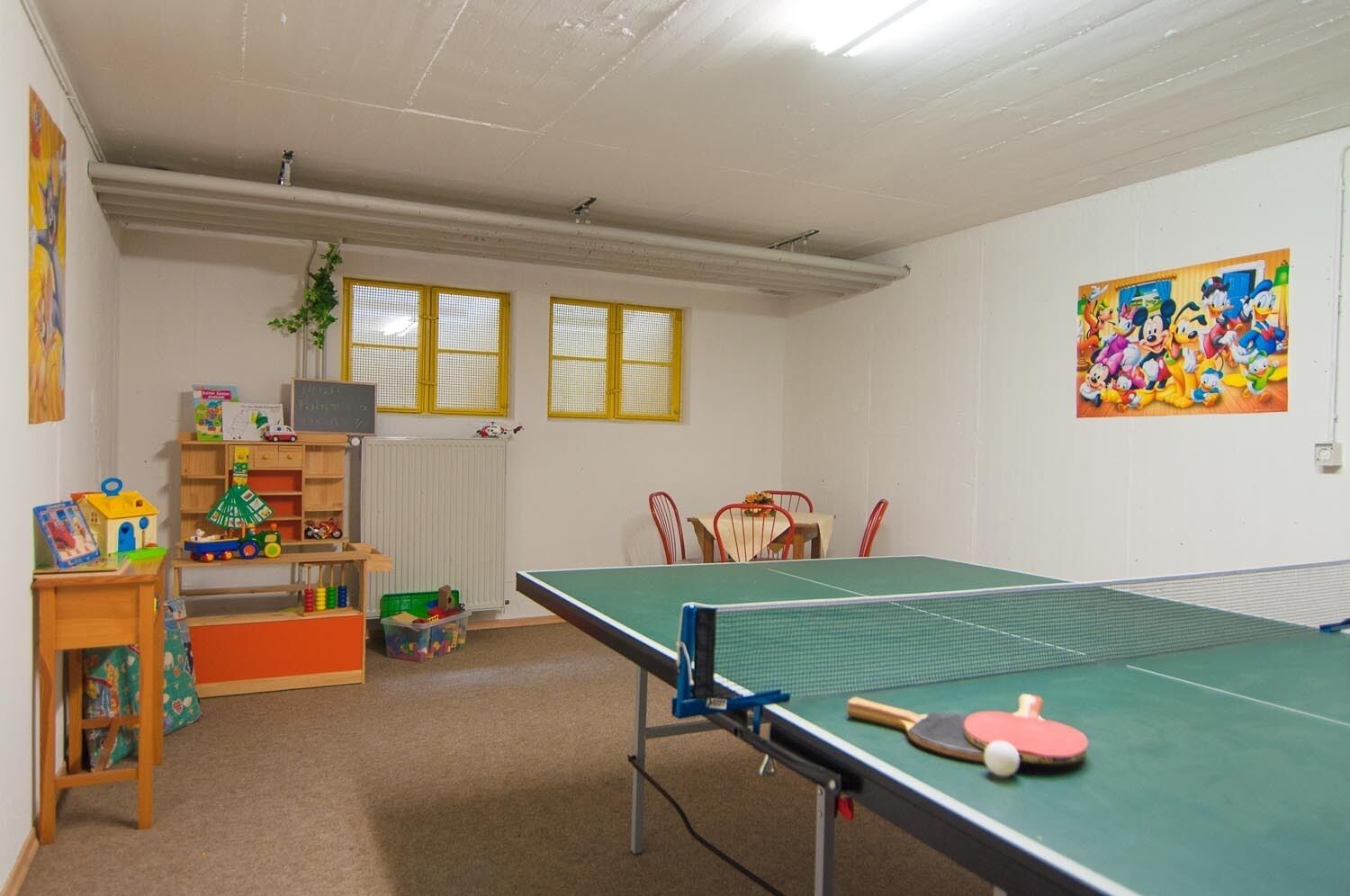 A room with a ping pong table, a bookshelf with toys, a radiator, and a window with yellow blinds.