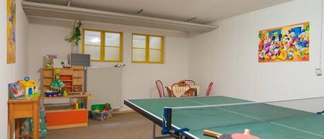 A room with a ping pong table, a bookshelf with toys, a radiator, and a window with yellow blinds.