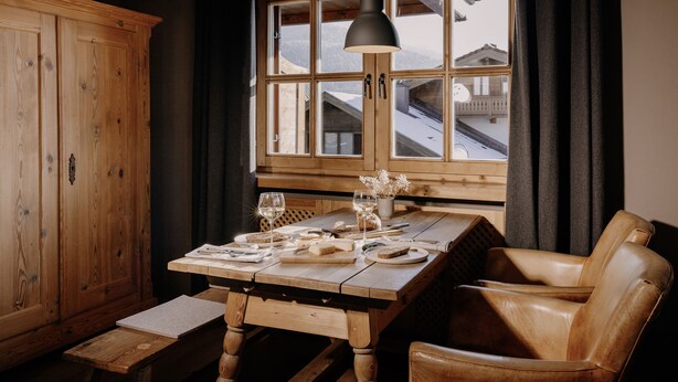 Una mesa de comedor de madera dispuesta con platos, vasos y cubiertos, una silla de cuero, y una ventana con vistas a edificios.