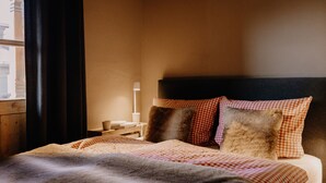 Una cama bien hecha con una colcha y almohadas a cuadros, una lámpara de noche y vistas a un edificio.